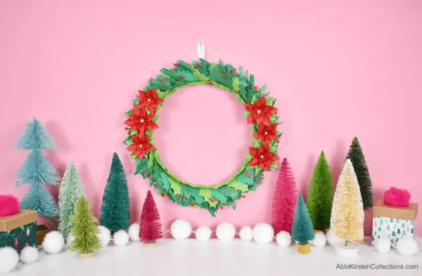 In front of a pink wall, on a white table, sit colorful Christmas trees, a string of white baubles, and small wrapped presents. A paper poinsettia and holly wreath hangs on the wall.