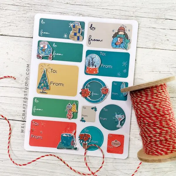 A sheet of colorful DIY wintery Christmas gift tags next to a spool of red and tan striped string sitting on a white wooden table. 