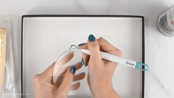 An image shows Abbi's hands, holding an acrylic dick ornament and a Cricut weeding tool, removing the plastic film from the ornament.