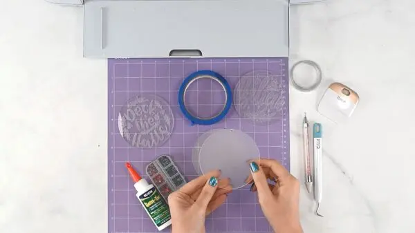 The supplies needed to make engraved acrylic Christmas ornaments are laid out on a purple cutting mat. You'll need acrylic disks, Cricut engraving tools, glue, some gems for decoration, and a Cricut cutting machine.