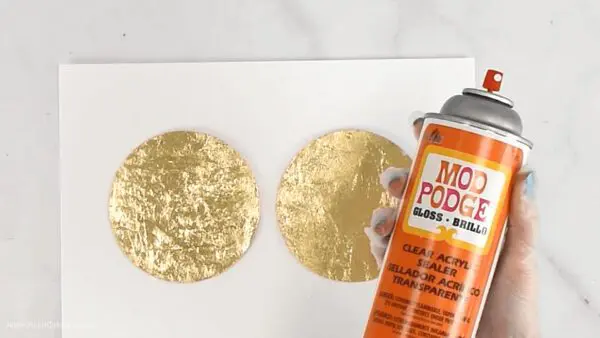 Abbi holds a can of Mod Podge pray next to two gilded gold acrylic ornaments.