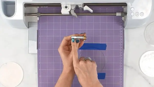 Abbi holds an engraving tip for the Circut maker in her hands, over a purple cutting mat with an acrylic ornament disk taped down, sitting inside a Cricut cutting machine.