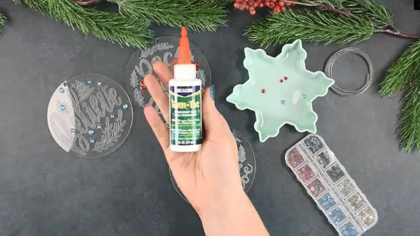 A white hand is pictured holding a bottle of Gemtac glue. Engraved Christmas ornaments and a tray of colorful gems sit laid out on a black tabletop.