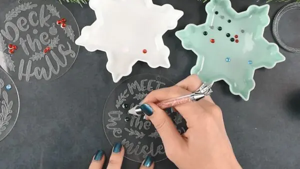 Abbi's hand is pictured using a gemstone pen to apply small gemstones to engraved acrylic Christmas ornaments. The small gems sit in white and green snowflake-shaped dishes.