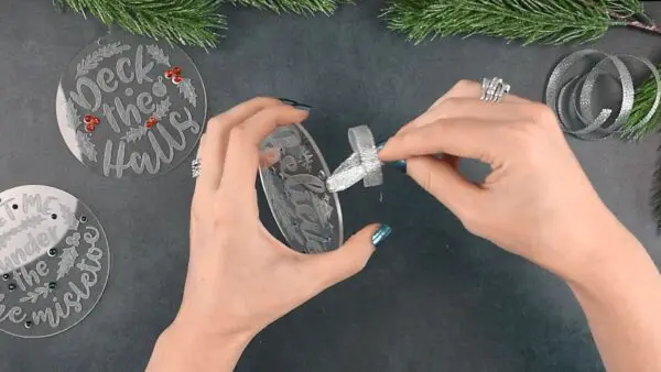 Abbi's hands are pictured adding a length of silver ribbon to the top of an engraved acrylic Christmas ornament.