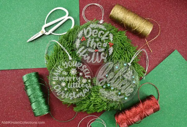 Three engraved acrylic Christmas ornaments sit on a Christmas wreath surrounded by red, green, and gold craft supplies.