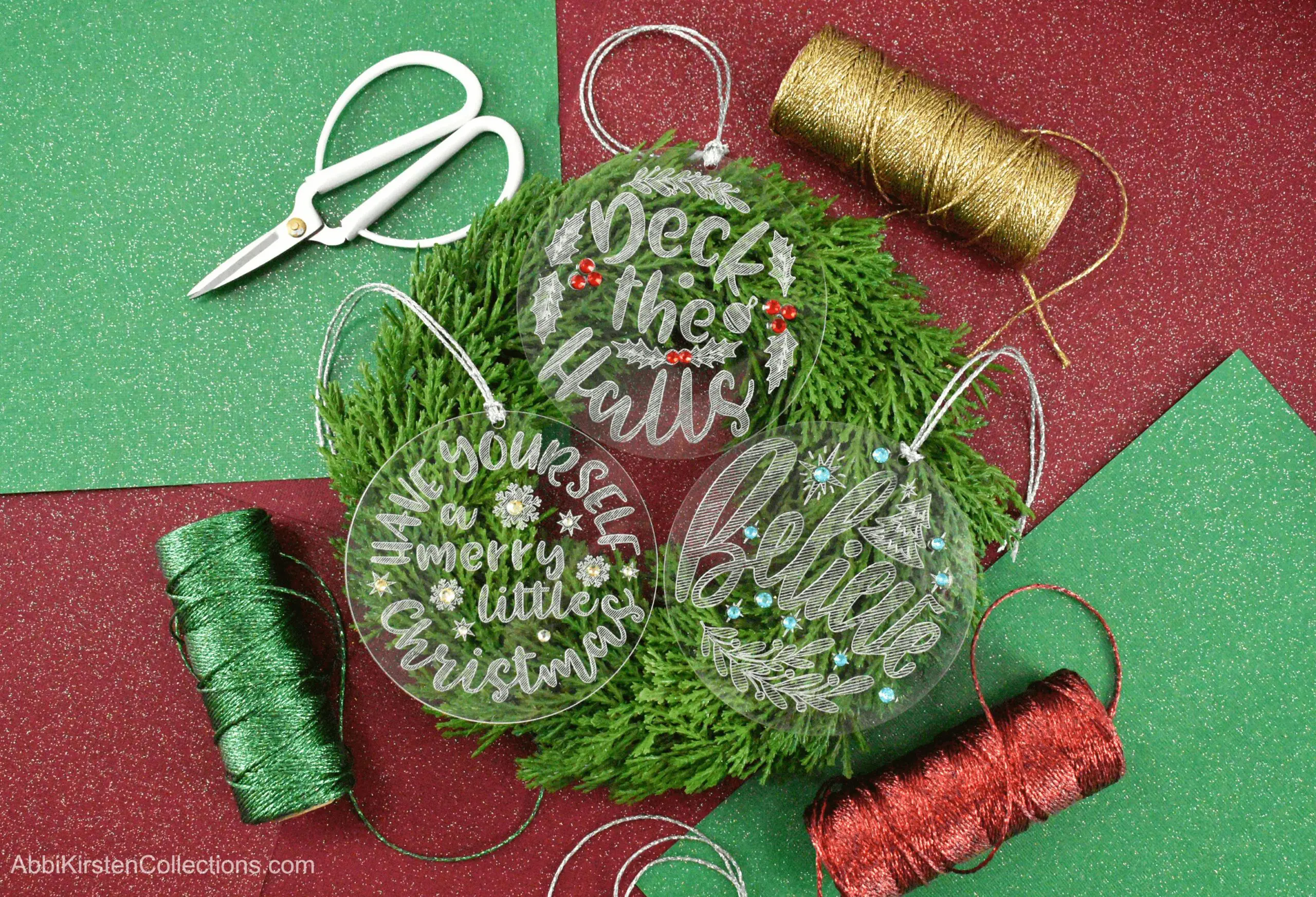Three engraved acrylic Christmas ornaments sit on a Christmas wreath surrounded by red, green, and gold craft supplies.
