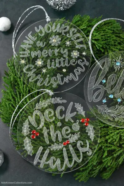 Three completed engraved acrylic Christmas ornaments sit on top of a Christmas wreath. Each ornament is engraved with a classic Christmas saying and decorated with gemstones and silver ribbons.