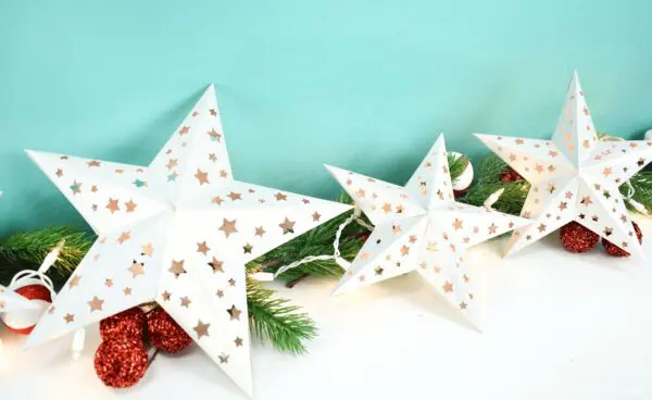Three white paper stars in varying sizes attached by a string of white lights. The star garland is laying on festive pine branches.