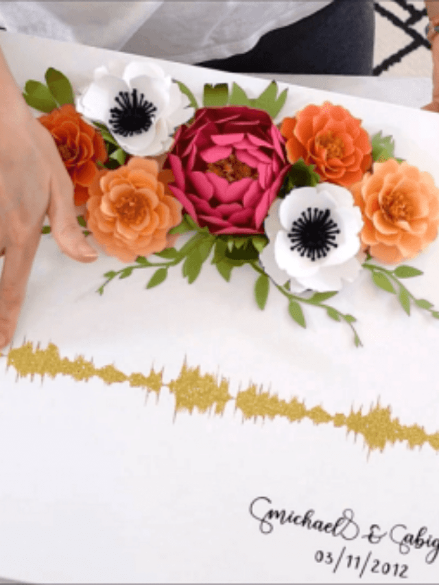 A person pressing down a gold printed soundwave on a white canvas decorated with paper peonies, roses and poppies with some green paper leaves.