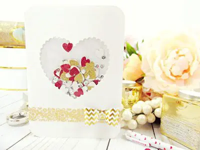 A white paper card with a gold and white bow tied near the bottom. The card has a heart-shaped window filled with gold, white and red paper heart confetti. 