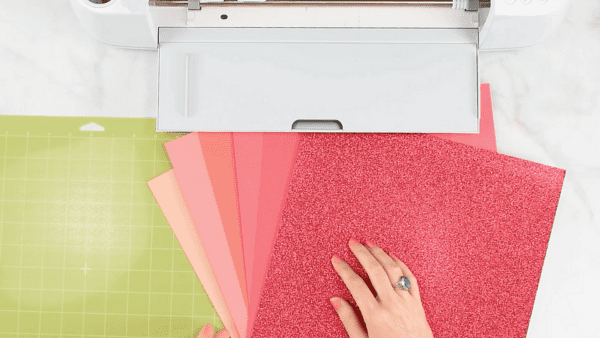 A variety of pink-hued cardstock paper on an avocado green mat next to a white Cricut Maker.