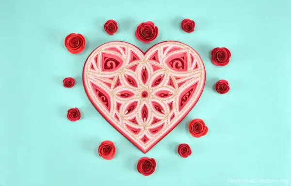 A red, pink and glitter heart mandala surrounded by red paper roses on a blue table.