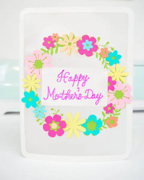 A white Happy Mother's Day card with a colorful floral wreath set on a white desk.