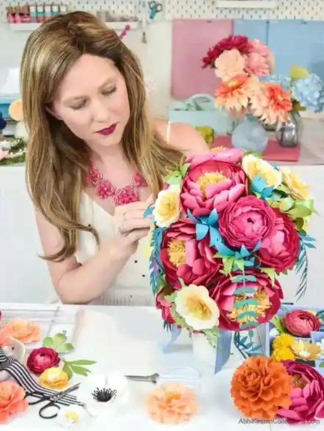 A woman wearing a sleeveless white dress with a flower necklace, arranging a cascading paper flower bouquet made of pink paper peonies and yellow buttercups.