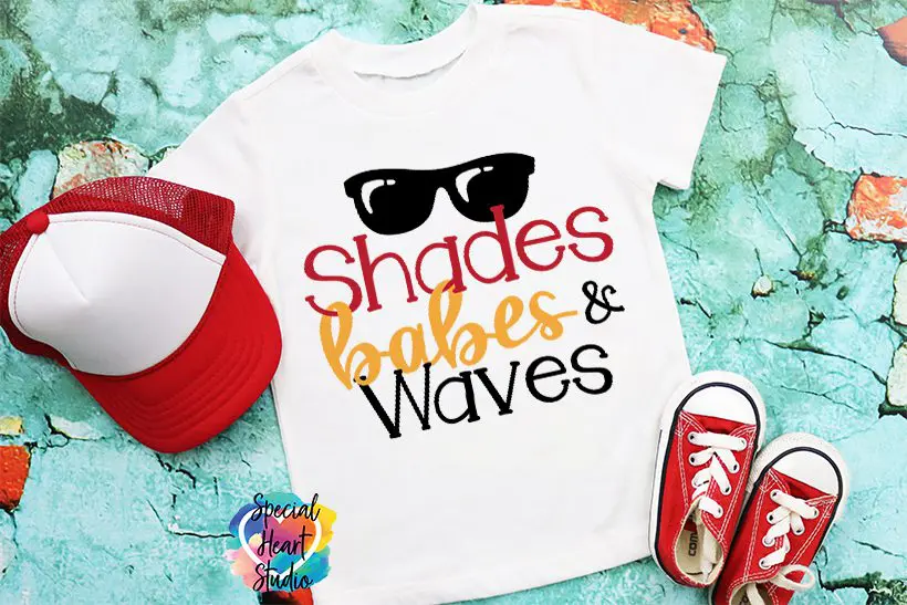A t-shirt with a design that says "Shades, Babes, and Waves" and a sunglasses illustration lays on a green floor with red Converse shoes and a red and white hat.