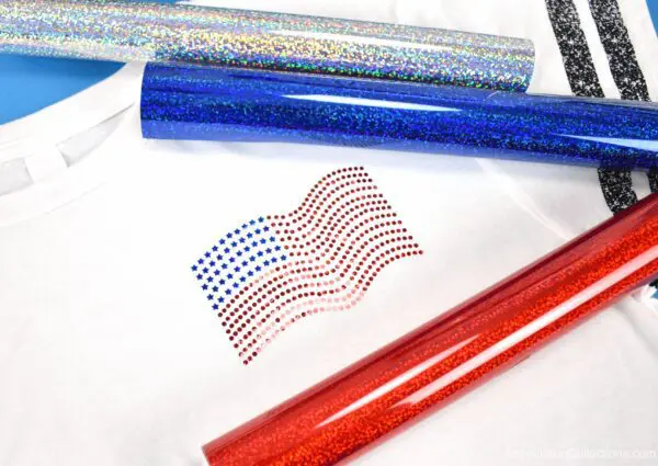 A faux rhinestone American flag made with holographic sparkle iron-on vinyl. on the pocket of a white t-shirt. The design is surrounded by rolls of Cricut holographic heat transfer vinyl.