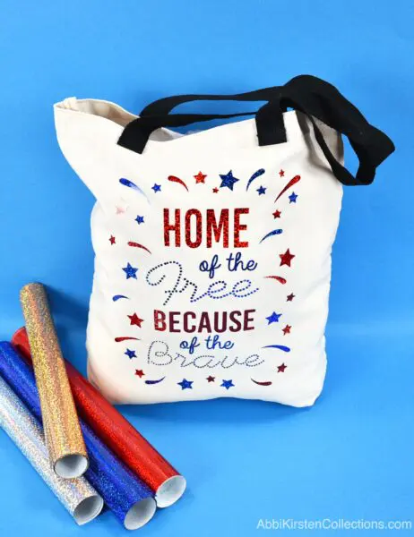 A cream-colored tote bag and rolls of gold, blue, red, and silver iron-on vinyl lay on a blue background. The text on the white tote bag reads "Home of the free because of the brave" in holographic sparkle iron-on from an SVG design.