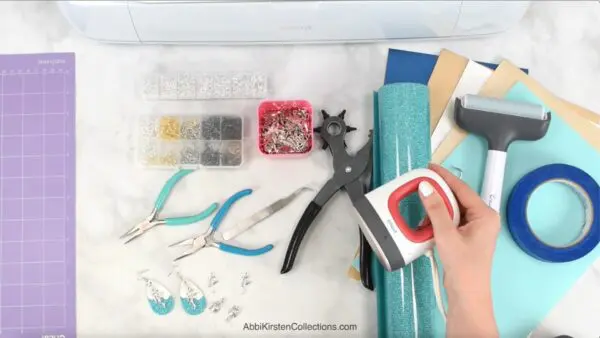 Supplies for making faux leather earrings next to a Cricut machine on a marble workspace. 