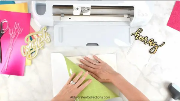 A pair of hands peeling a light green mat away from the back of a piece of paper. A white Cricut machine along with various tools lay nearby.