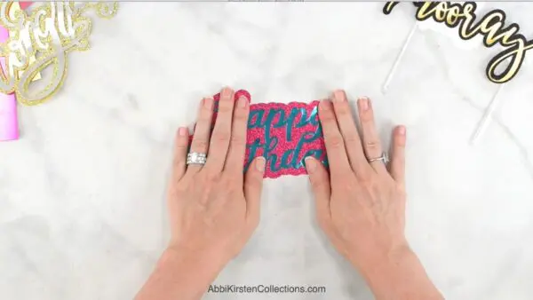 Two hands press down a blue happy birthday cutout on top of hot pink glitter paper.