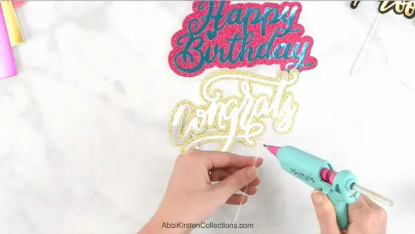 A pair of hands use a hot glue gun to place glue on plastic floral picks. Two cake toppers lay on a marble work surface.e