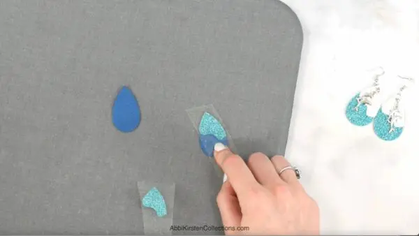 Blue and turquoise faux leather pieces being aligned and pressed together on a grey mat next to an assembled pair of white and turquoise faux leather earrings. 