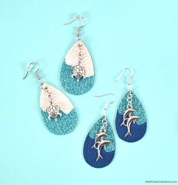 Two sets of teardrop-shaped faux leather earrings, one white and turquoise with silver sea turtle charms and the other turquoise and blue with silver dolphin charms, sitting on a light blue table. 
