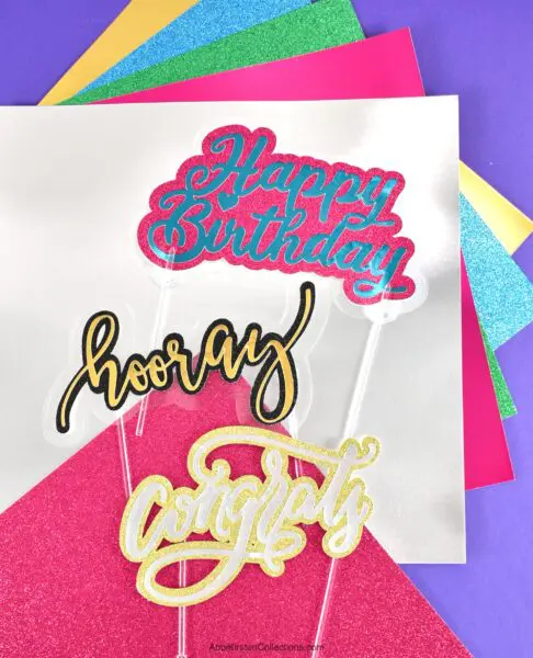 Three multicolored cake toppers that read "happy birthday", "hooray" and "congrats" lay on top of sheets of various colored paper.