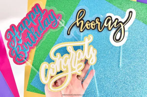 Three colorful cake toppers that read "congrats", "hooray", and "happy birthday" lay on top of sheets of glittery multicolored paper.