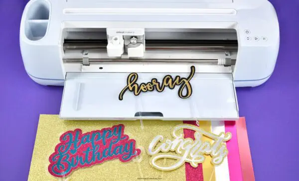 Three different colorful signs that read, "hooray", "happy birthday", and "congrats" laying on a white Cricut and multicolored pieces of paper.