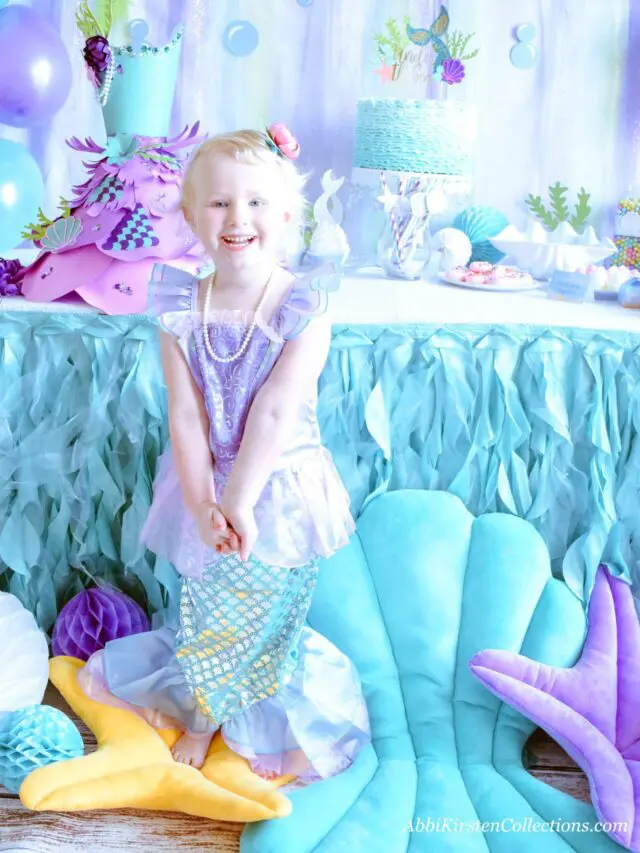 Ella is surrounded by Under the Sea themed props and decor in blues and purples. There are stuffed shells to sit on, a wavy blue tablecloth, and mermaid treats.