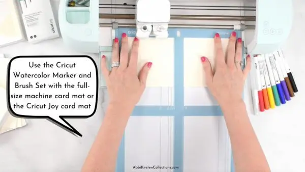 A close-up to Abbi Kirsten's arms loading the Cricut watercolor cards on a blue mat into a Cricut machine. A voice bubble says "Use the Cricut Watercolor Marker and Brush Set with the full-size machine card mat or the Cricut Joy mat!"