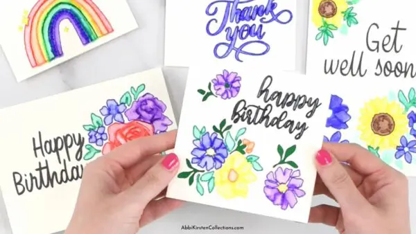 Abbi Kirsten holds a white watercolor "Happy Birthday" card above other watercolor cards that say "Get Well Soon" and "Thank You.' Cards are decorated with flowers and a rainbow. 