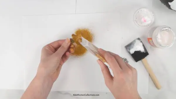 A woman uses mod podge to apply gold glitter to the stem of a craft pumpkin.