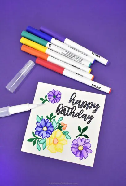 On a dark blue background lies Cricut watercolor pens and a handmade birthday card. The white card says "Happy Birthday" and has decorated flowers and leaves drawn on. 