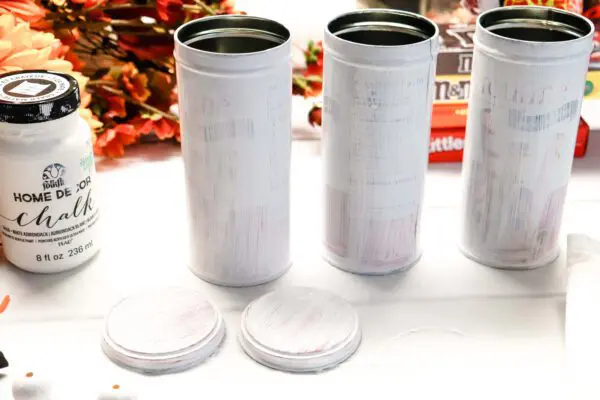 Amid fall faux flowers, three tall cookie tins are drying after a coat of white chalk paint. The bottle of the white paint is to the left of the tins. 