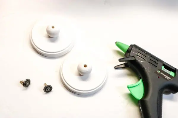 A close-up of a glue gun laying next to painted tin can lids with painted wooden beads on top and fasteners on the left. The tin can lids are part of the Halloween trick or treat craft using repurposed cookie tins. 