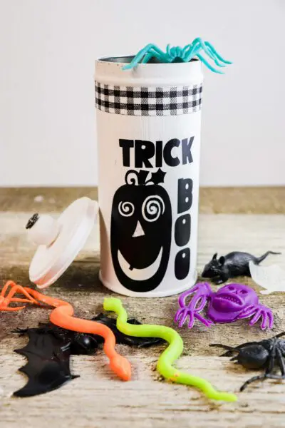 A repurposed cookie tin has been painted white. Black vinyl words "trick or treat" and a pumpkin have been added, as well a buffalo black and white ribbon encircling the top. The lid rests against the tin, which is surrounded by plastic creepy crawlies. 