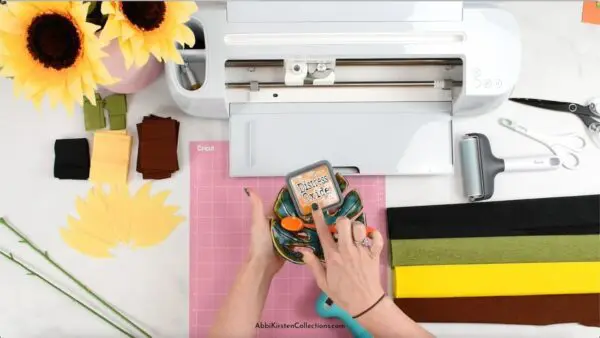 Abbi Kirsten's hands hold distress ink and an application brush, over a Cricut machine and fabric mat, floral wire and crepe paper sunflowers and additional sunflower elements.