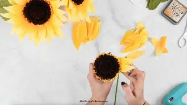 An overhead view of Abbi Kirsten's hands adding petals to the crepe paper sunflower center. A couple of finished crepe paper sunflowers sit at the top of the work table.