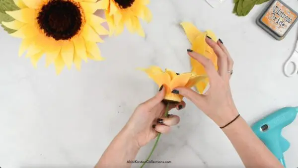 Abbi's thumb gently presses a yellow paper petal to the center of the crepe paper sunflower center. A glue gun, scissors, distressed ink and finished sunflower blooms dot the work table.