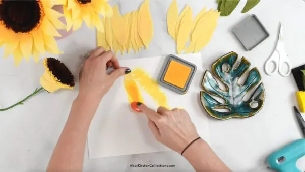 Amid finished crepe paper sunflowers and cut-out petals and supplies, Abbi Kirsten's hands add distress ink to the yellow crepe paper petals.