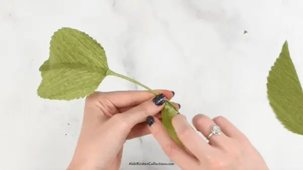 A close-up overhead view of Abbi Kirsten's hand gluing crepe paper green leaves onto a floral wire. An additional leaf lays off to the side.
