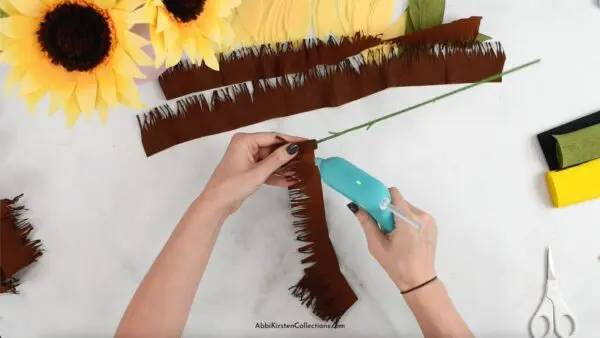 Abbi Kirsten's hands create the fringe flower center piece using brown strips of crepe paper and a hot glue gun. Rolls of crepe paper and paper sunflowers surround the table.