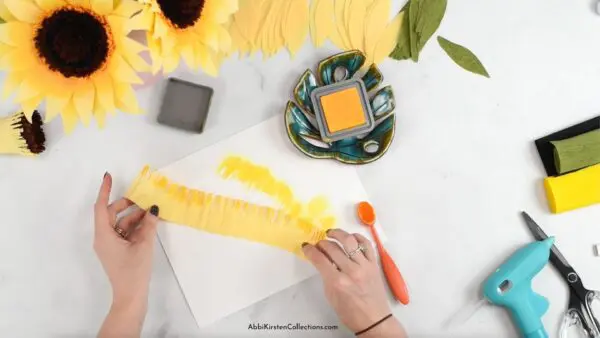 Abbi's hands hold a strip of yellow crepe paper that has distress ink applied to create a dimensional element. Supplies for the paper flower craft surround the active area.