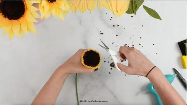 Abbi Kirsten's hands hold the crepe paper sunflower center while using detail scissors to trim the brown center. Bits of brown crepe paper freckle the work table.