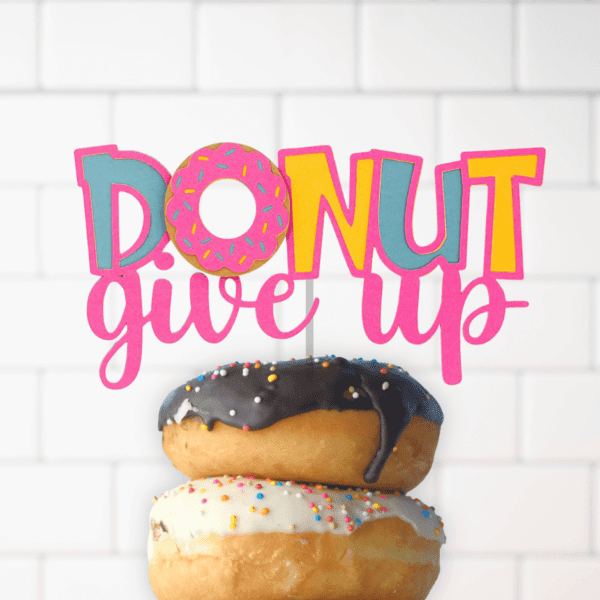 Two frosted donuts with sprinkles are stacked in front of a brick wall. A cake topper crowning the donuts says, "Donut give up." The colorful cut-out topper is a funny, free SVG file you can download.
