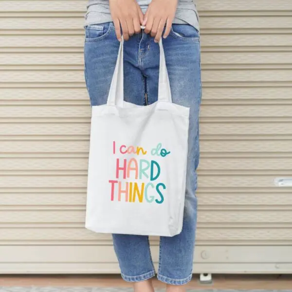 The "I can do hard things" free cut file was used to create the rainbow text on the beige totebag held by a woman against a garage door.