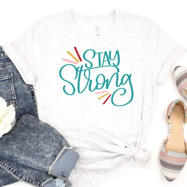 Jeans, slip-on shoes, and a white t-shirt that says "Stay Strong" lay on a white backdrop. This design is available as a free SVG cut file.
